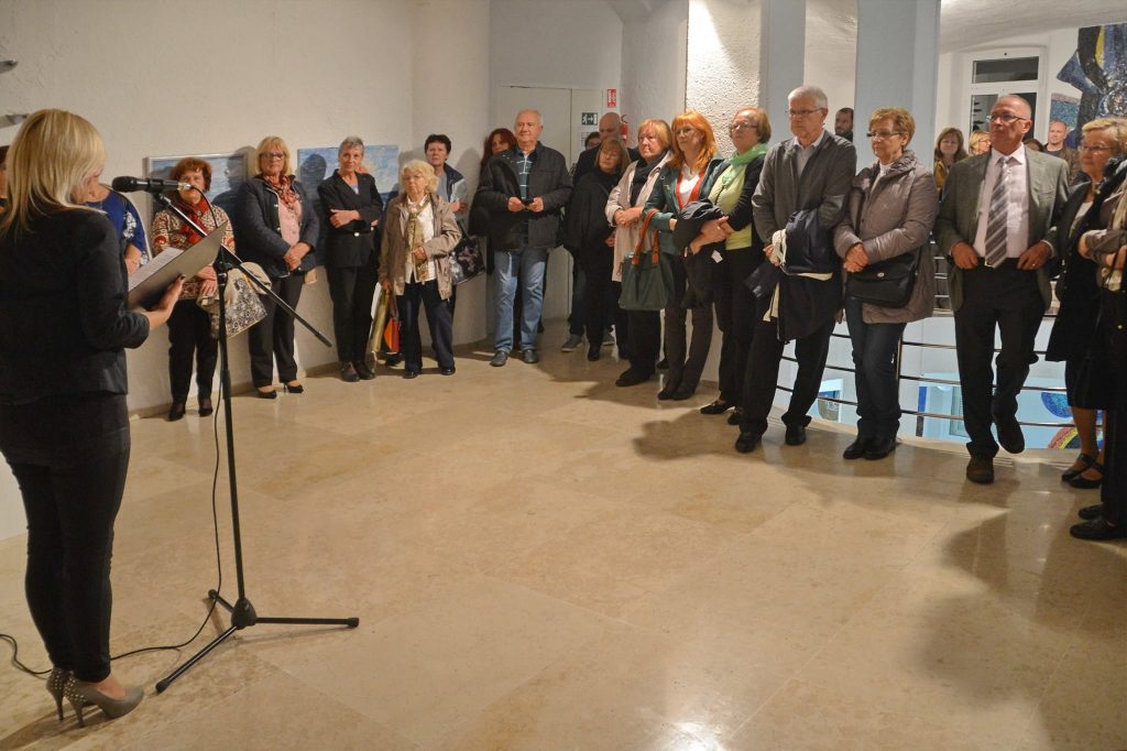 Dogodek. Ena gospa govori na mikrofon, na drugi strani gledalci.