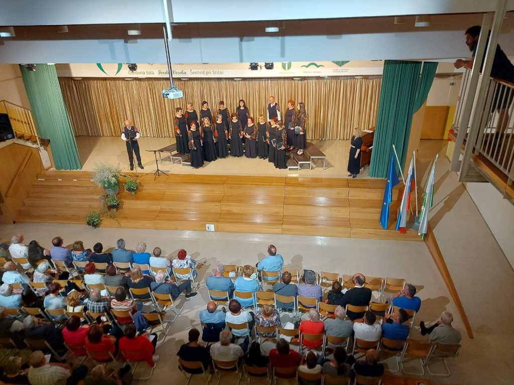 Pevski zbor med nastopom na odru. Spodaj na sliki je vidno občinstvo. Slika je posneta od zgoraj