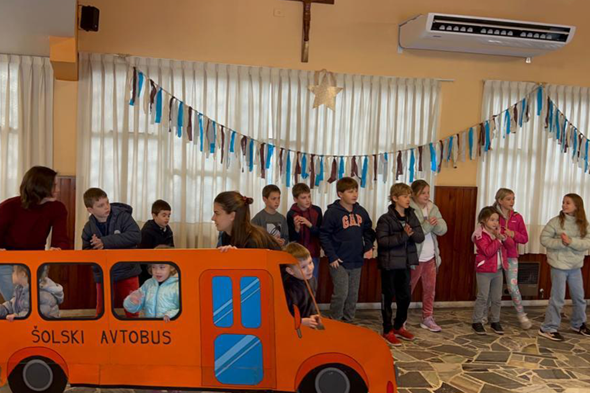 Otroci med šolskim nastopom. v ospredju je oranžen kartonast avtobus, ki je del predstave.