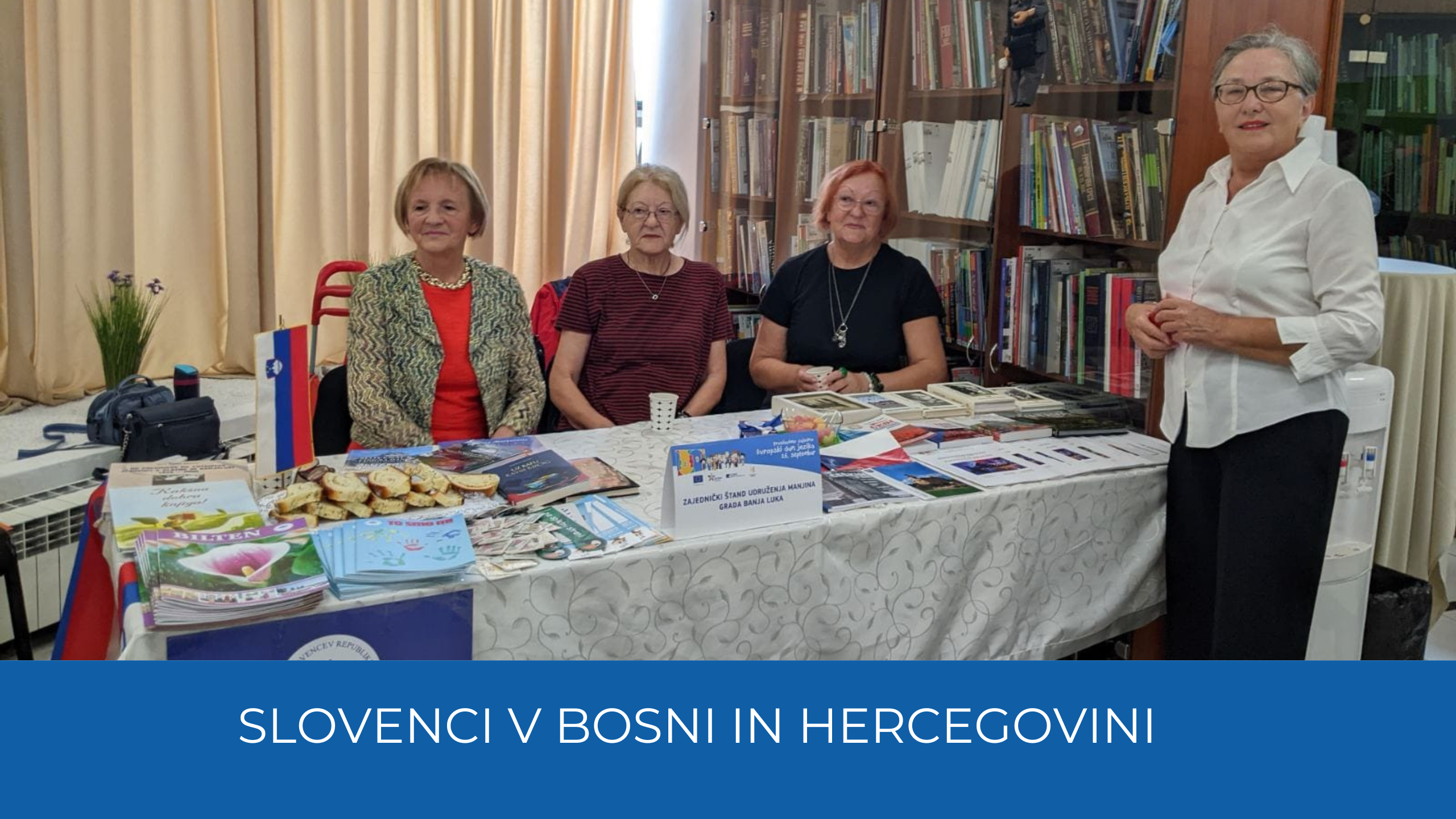 Ženske sedijo za stojnico s slovensko zastavo, letaki in potico