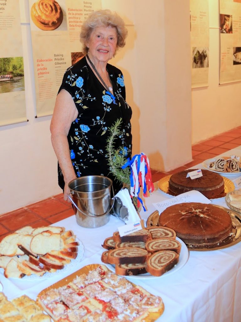 Starejša ženska stoji za mizo s slovenskimi dobrotami